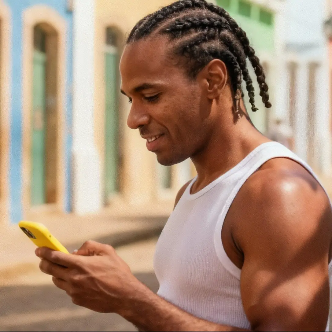 Homem sorridente com tranças usando um celular amarelo enquanto caminha em uma rua com casas coloridas. Representa a praticidade de usar o assistente financeiro Jota em qualquer lugar, direto pelo WhatsApp.