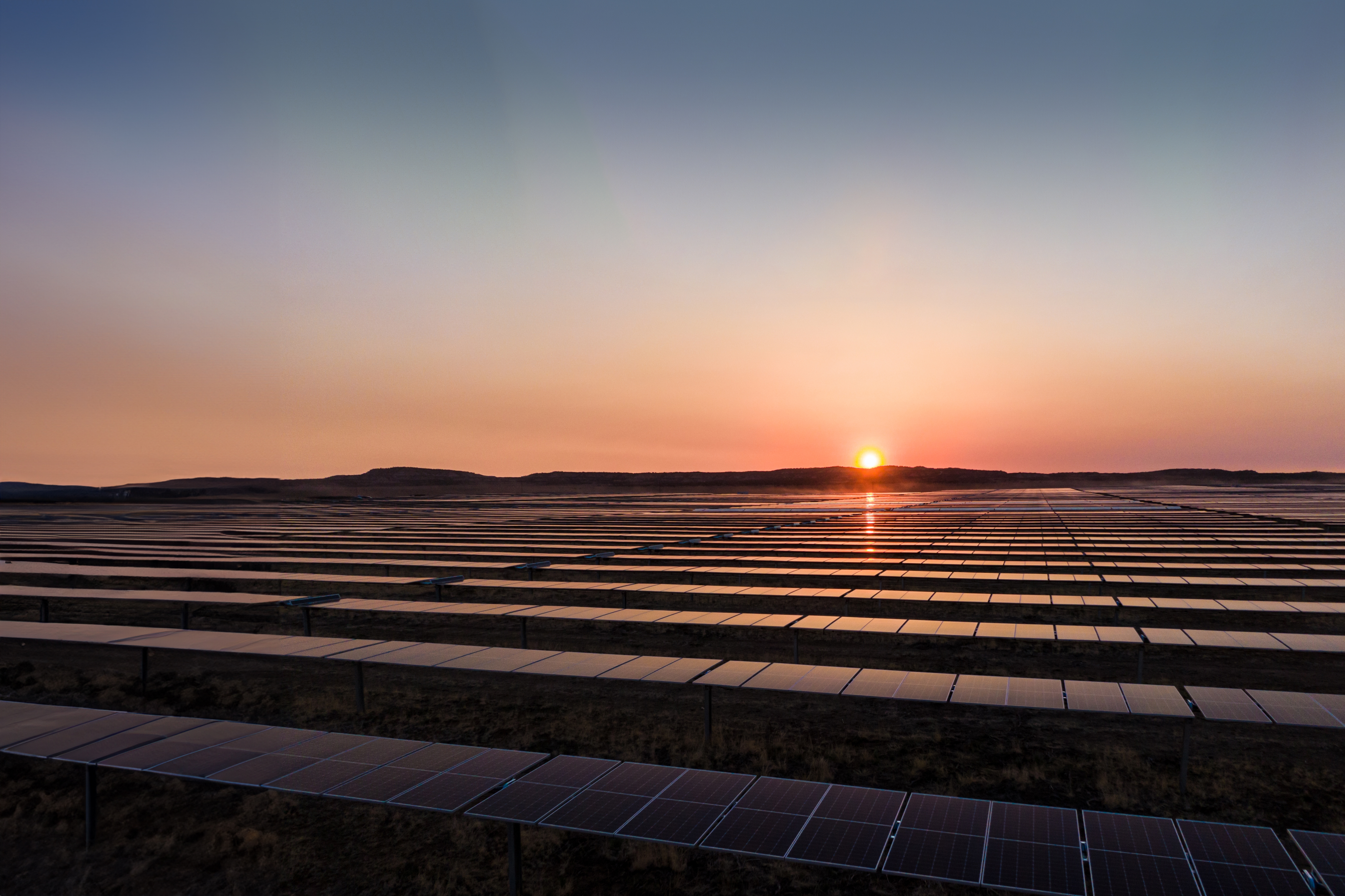 Vista panorâmica de uma grande fazenda solar com longas fileiras de painéis fotovoltaicos sob um pôr do sol alaranjado.