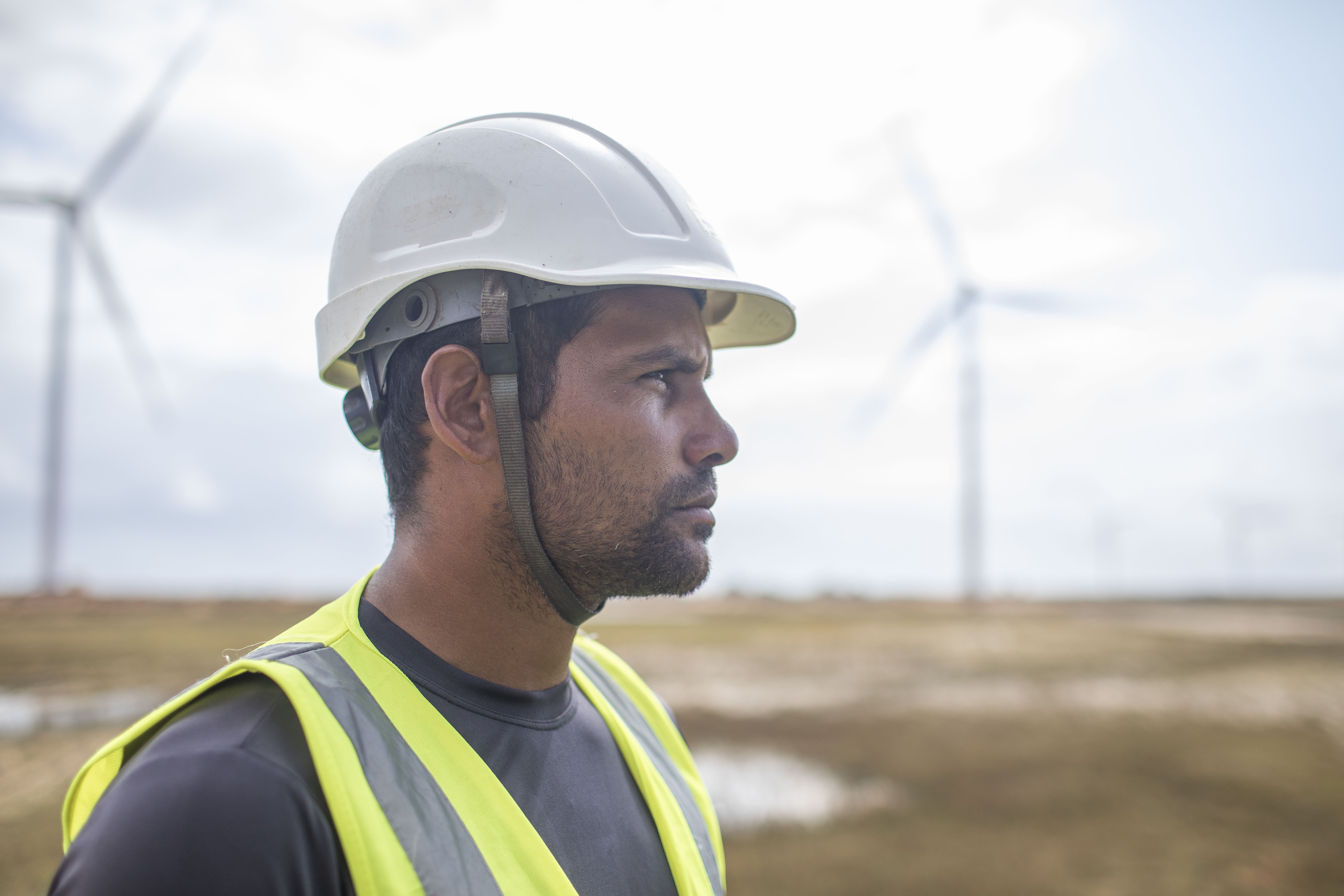 Perfil lateral de um trabalhador concentrado, usando capacete branco e colete de segurança amarelo de alta visibilidade, com aerogeradores desfocados ao fundo.