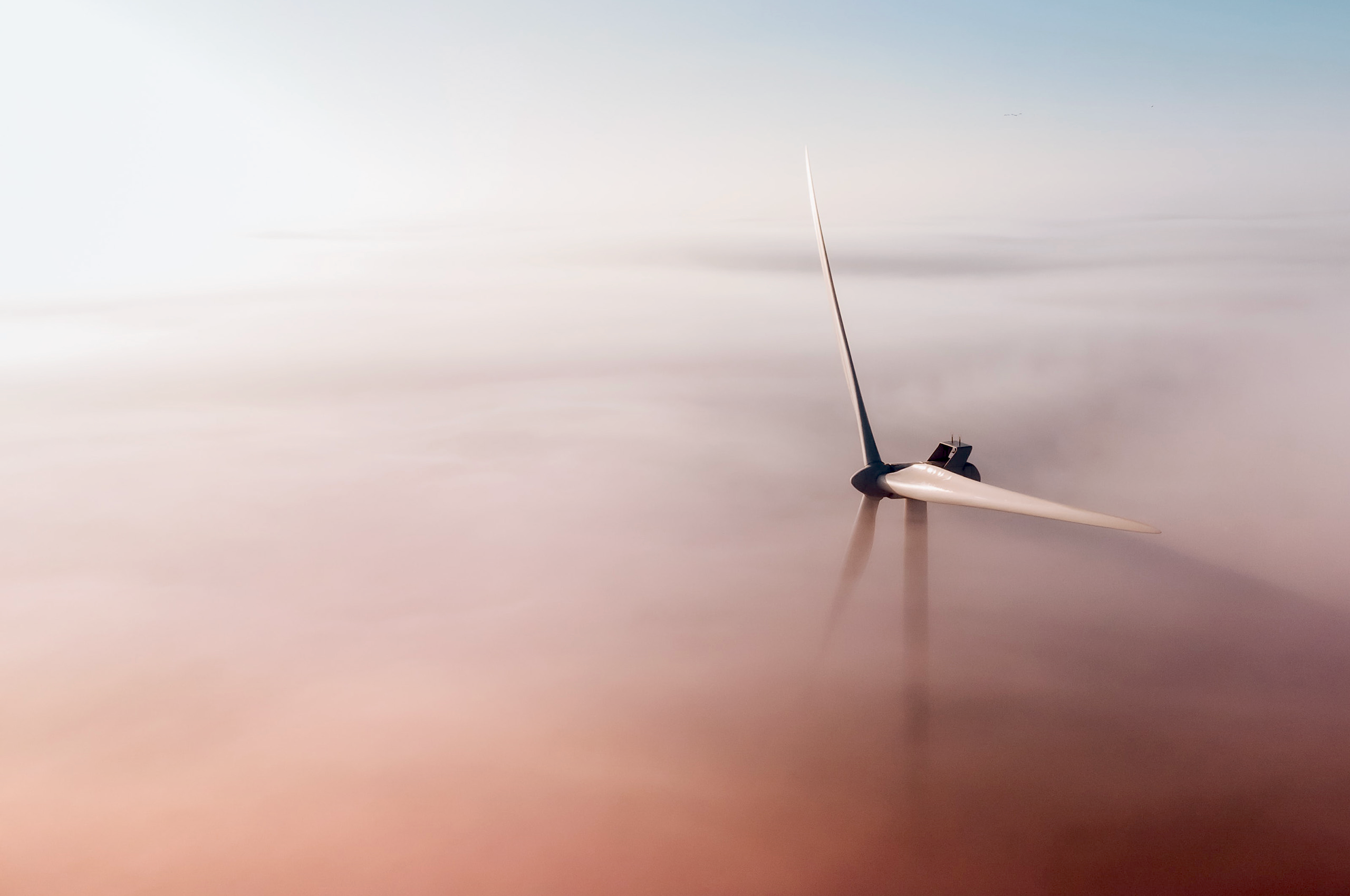 Um único aerogerador branco se destaca em meio a uma densa camada de neblina ou nuvens, iluminado pela luz suave do pôr do sol.