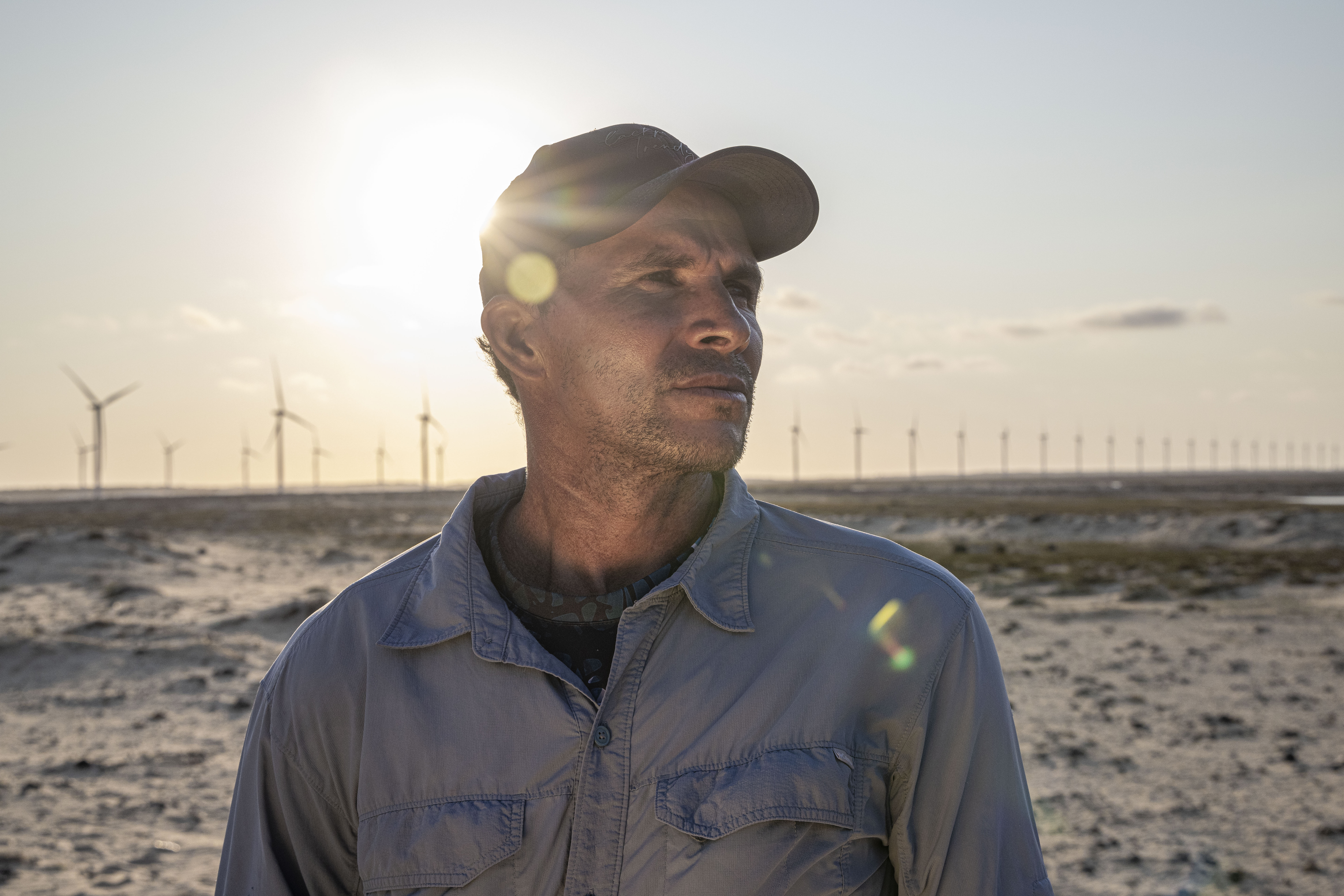 Perfil lateral de um trabalhador em um parque eólico, com o sol ao fundo e diversas turbinas eólicas estendendo-se pelo horizonte.