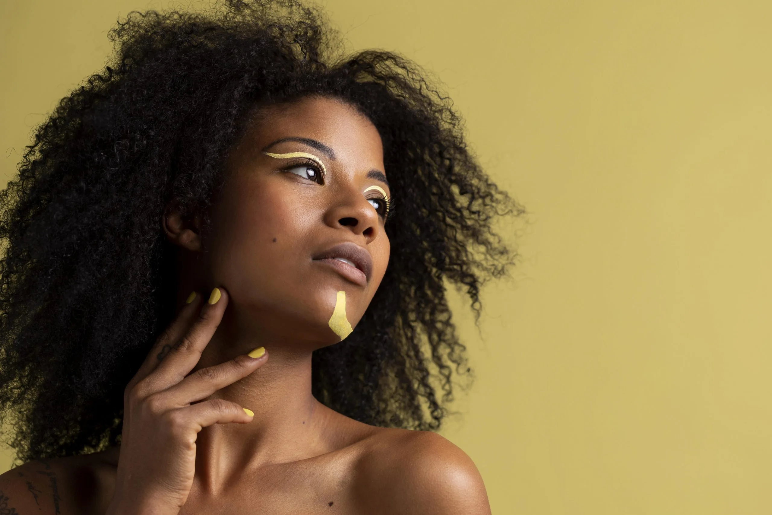 Mulher negra com maquiagem dourada nos olhos, a mão apoiada no pescoço, olhando para o lado.