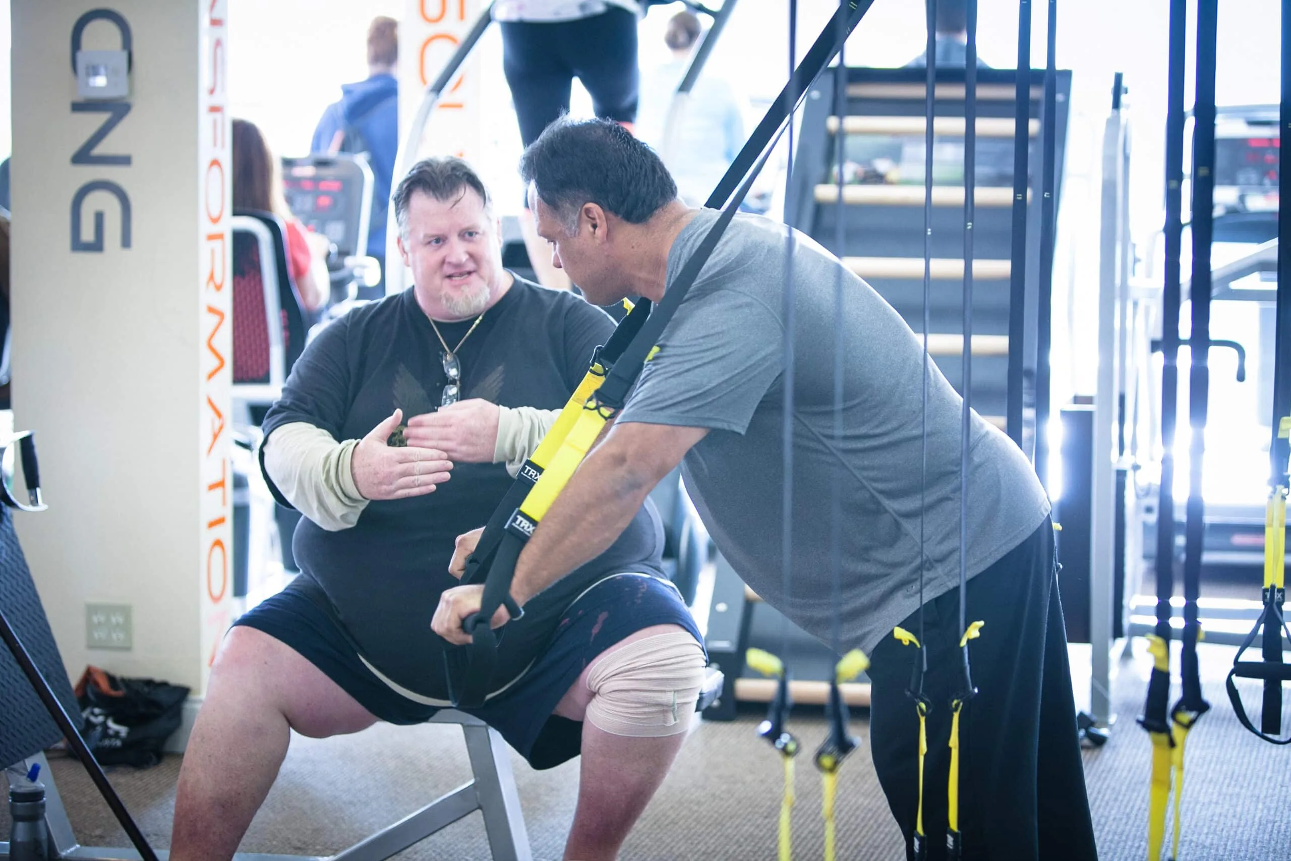 A trainer guides a client through suspension-strap training in a gym.