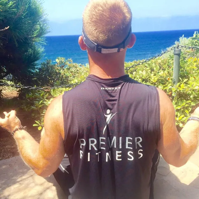 A person in a Premier Fitness tank top stands with arms open, facing the ocean from a coastal bluff.