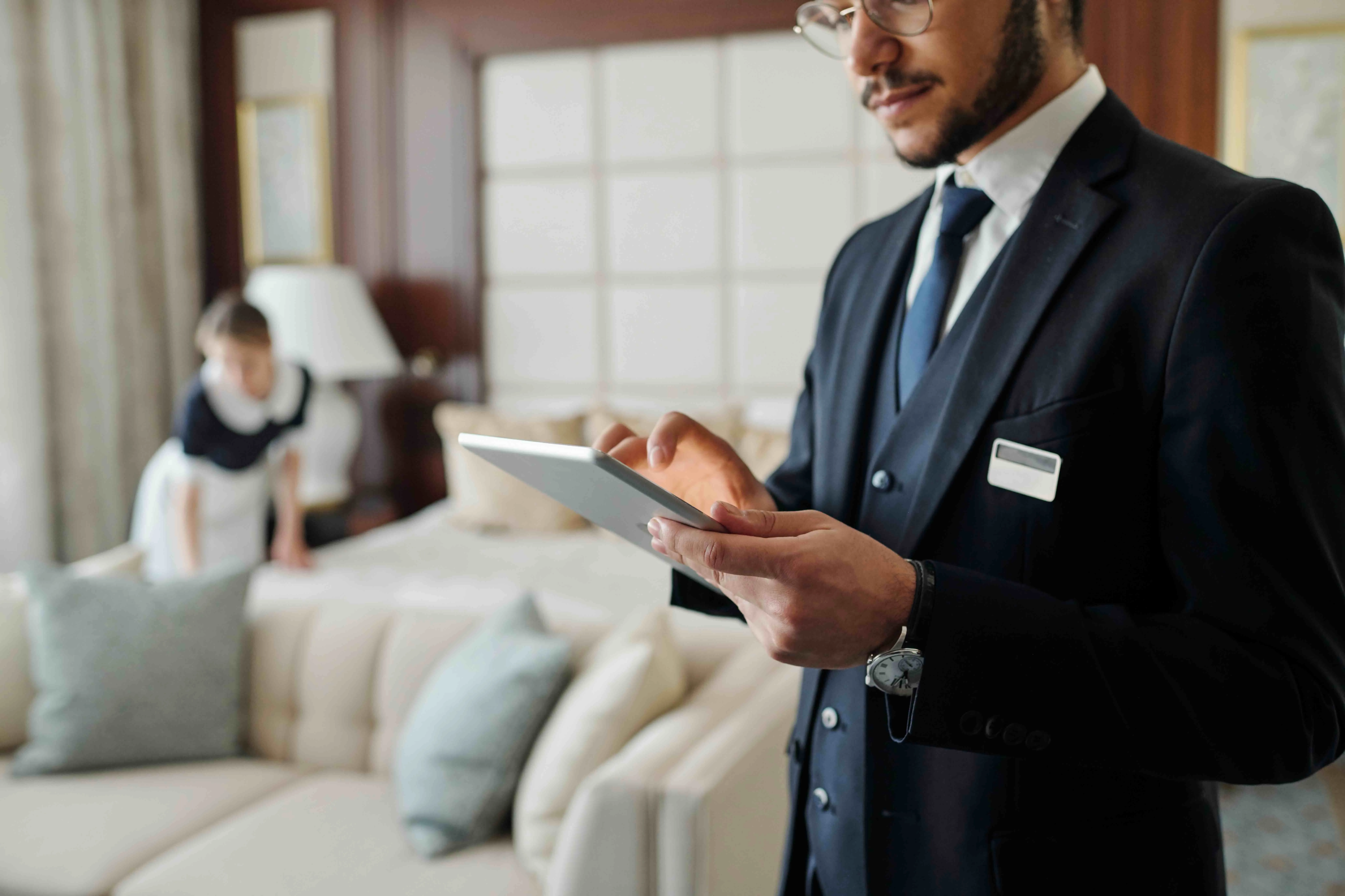 A hotel manager reviews a tablet in a guest room while a housekeeper makes the bed behind him.