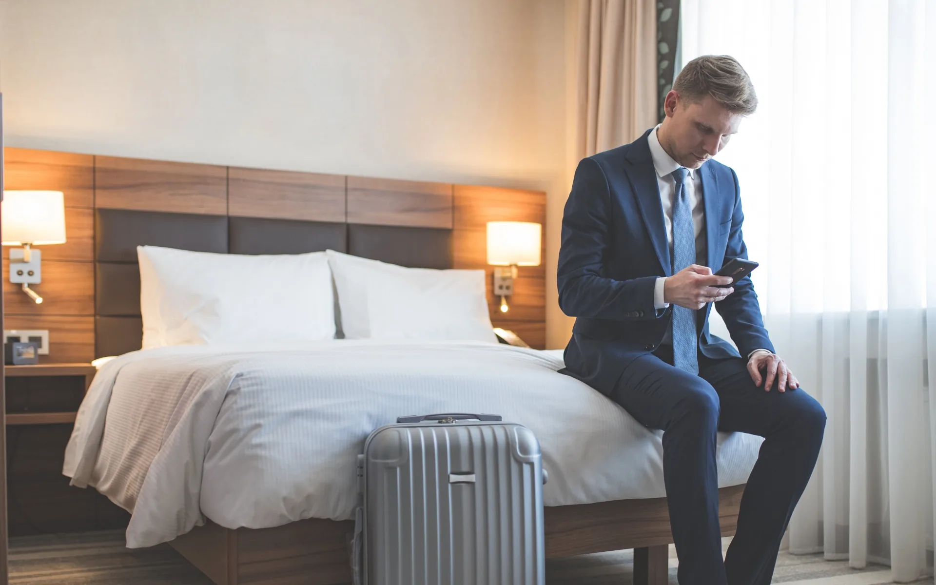 A business traveler sits on a hotel bed checking his phone, a suitcase beside him.