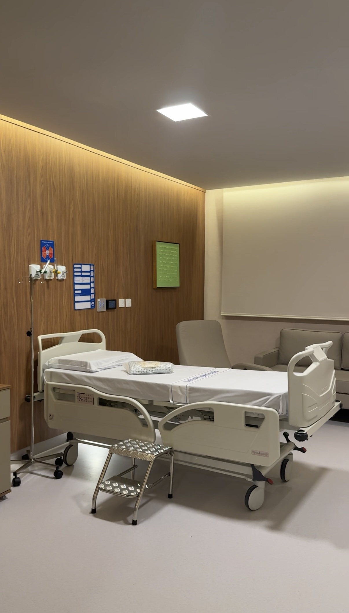 cama hospitalar elétrica, equipada com grades laterais de proteção e regulagem de altura e inclinação. Ao lado da cama há um suporte para soro (porta-soro) e um banco de apoio, utilizado para facilitar o acesso do paciente ao leito. Na cabeceira encontram-se os painéis de gases medicinais, tomadas elétricas, conexões para equipamentos hospitalares e campainha de chamada da enfermagem.