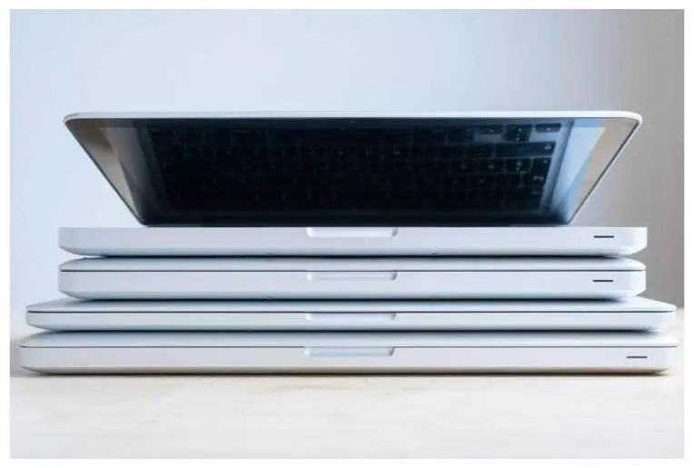 A stack of four silver laptops on a light wooden surface.