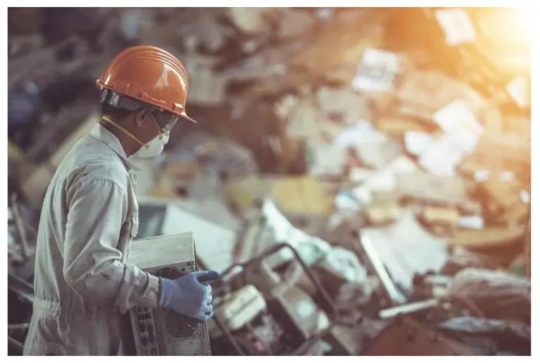A worker in a hard hat and respirator carries a device at an electronics recycling facility.
