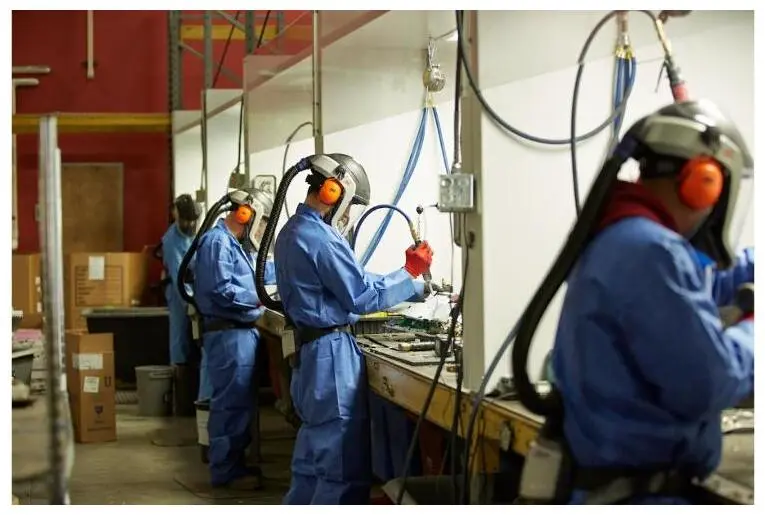 Workers in blue coveralls and respirators process electronics along an industrial line.