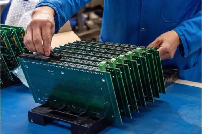 A worker in a blue smock handles a row of green printed circuit boards.