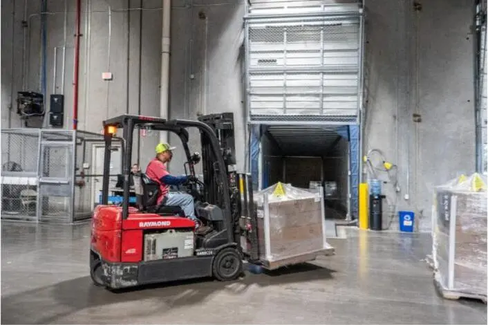 A forklift loads a shrink-wrapped pallet into a trailer at a warehouse dock.