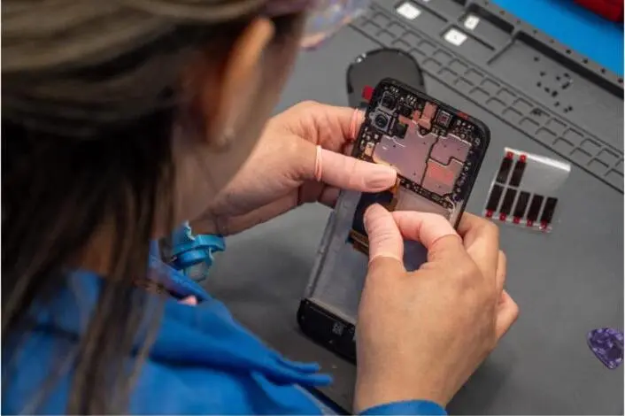 A technician in safety glasses works on the exposed board of a mobile device.