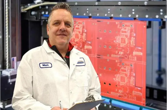 An engineer in a lab coat holds a clipboard beside a large red PCB panel.