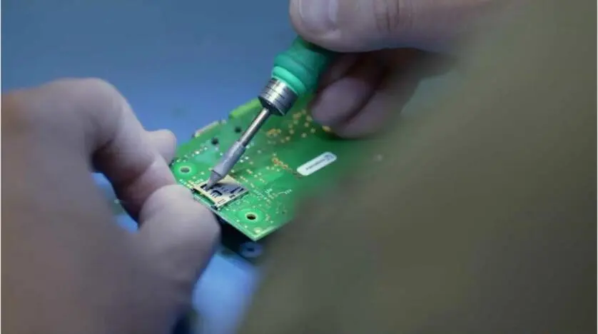 A technician's hands using a soldering iron on a green circuit board.