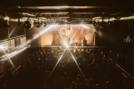 A live music venue crowd facing a brightly lit stage.