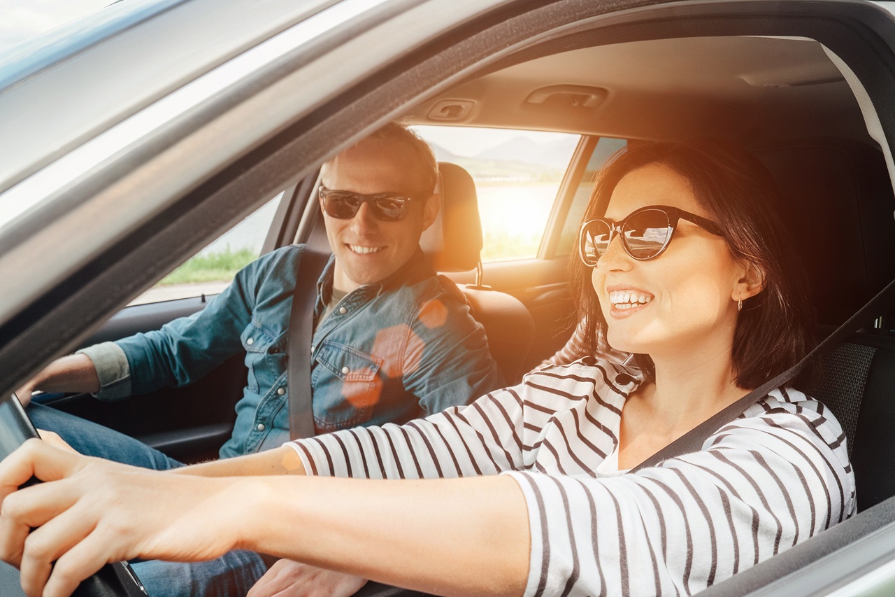 Casal sorrindo dentro do carro ao pôr do sol, com a mulher ao volante.
