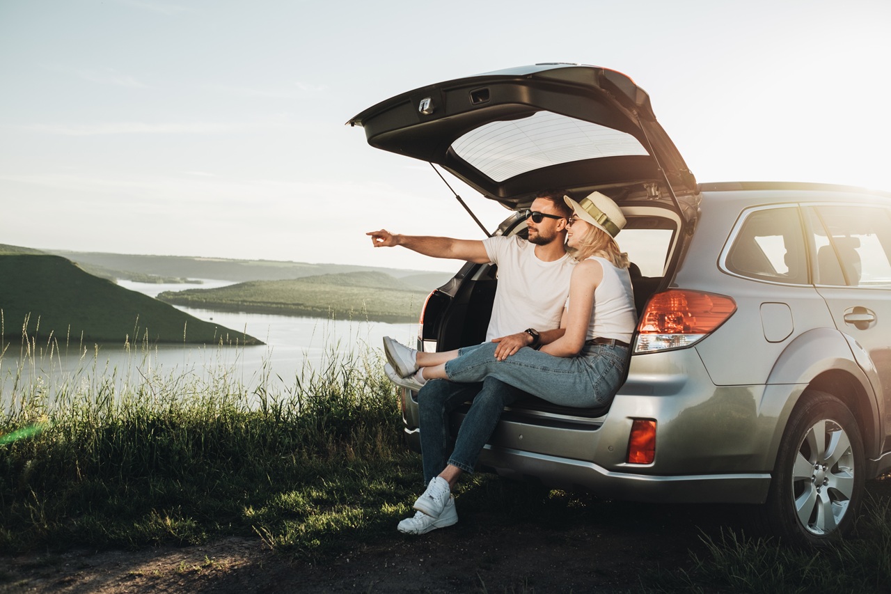 Casal sentado no porta-malas aberto de um SUV, diante de uma paisagem com lago.