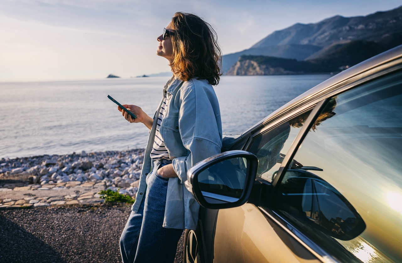 Mulher em pé ao lado do carro, à beira-mar, olhando o celular.