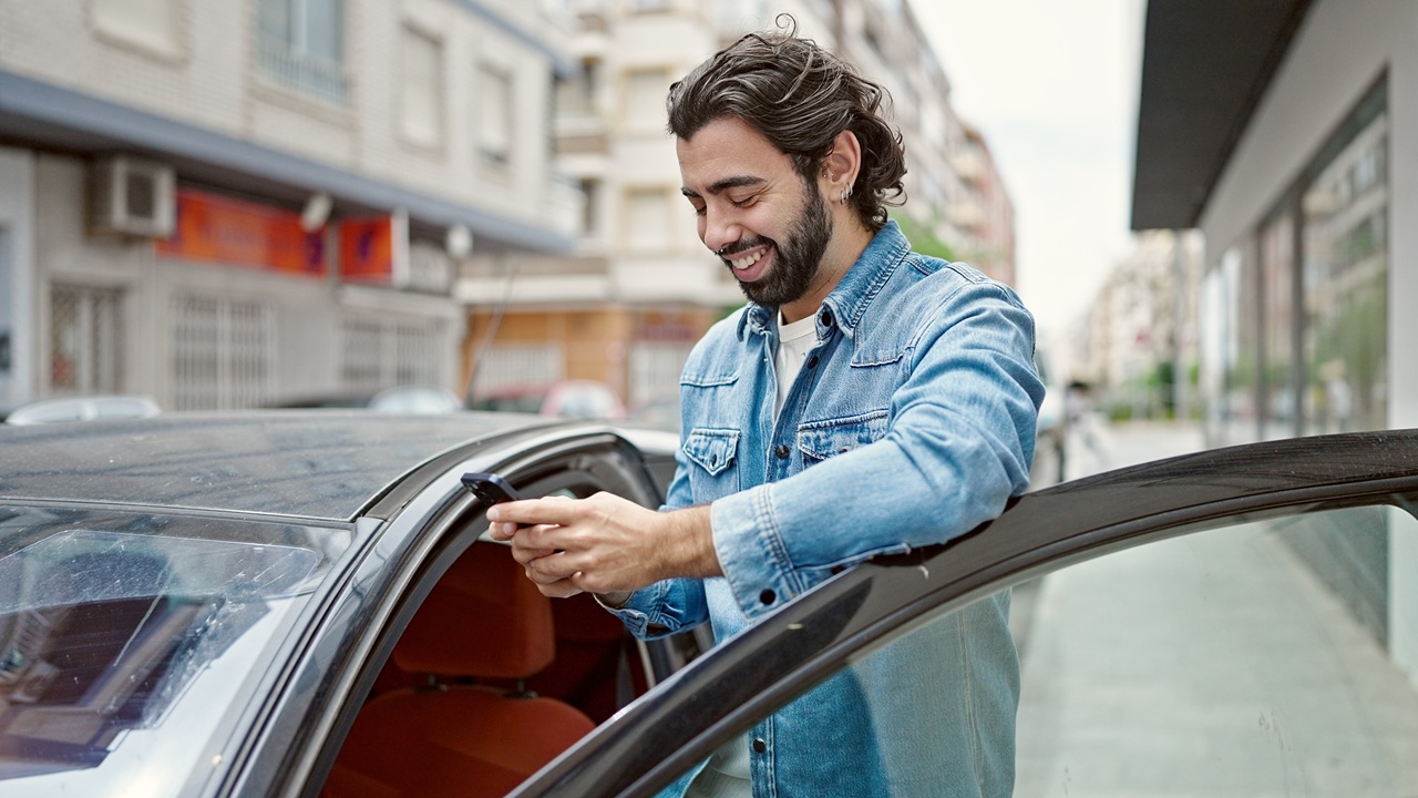 Homem de jaqueta jeans ao lado do carro, na rua, mexendo no celular.