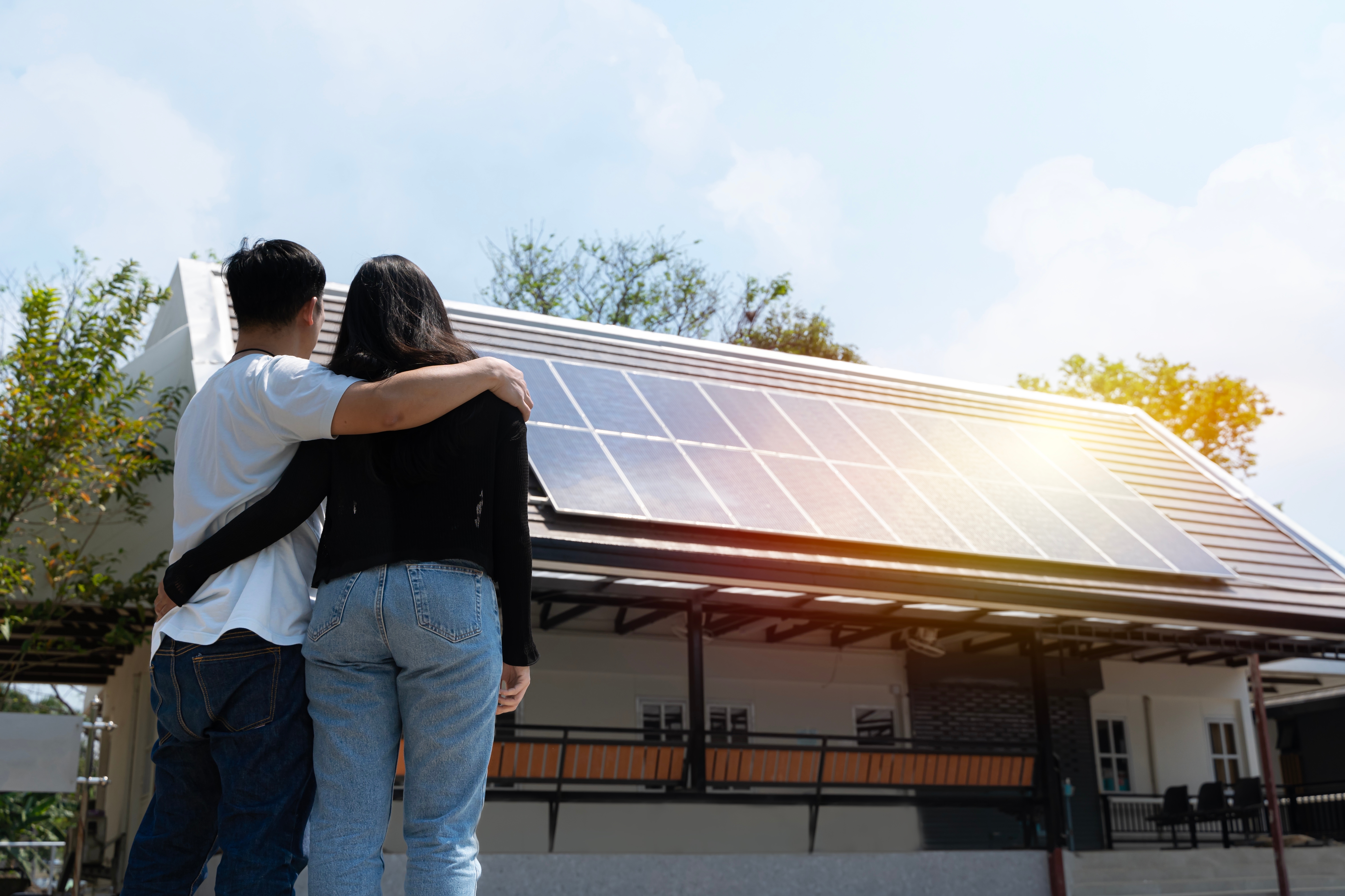 Casal abraçado, de costas, observando uma casa com painéis solares ao pôr do sol.
